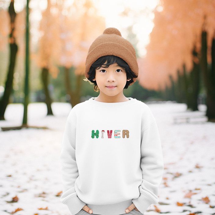 Smiling child wearing a white sweatshirt with colorful “HIVER” letters, festive winter design by KOULEURS.
