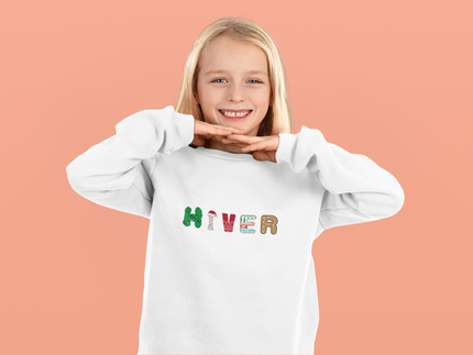 Smiling child wearing a white sweatshirt with colorful “HIVER” letters, festive winter design by KOULEURS.